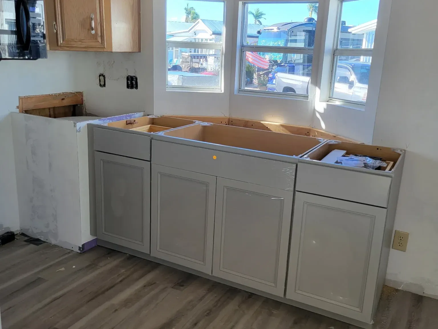 New gray shaker base cabinets set and leveled under a bright bay window, tops open ahead of the countertop template, new plank flooring down