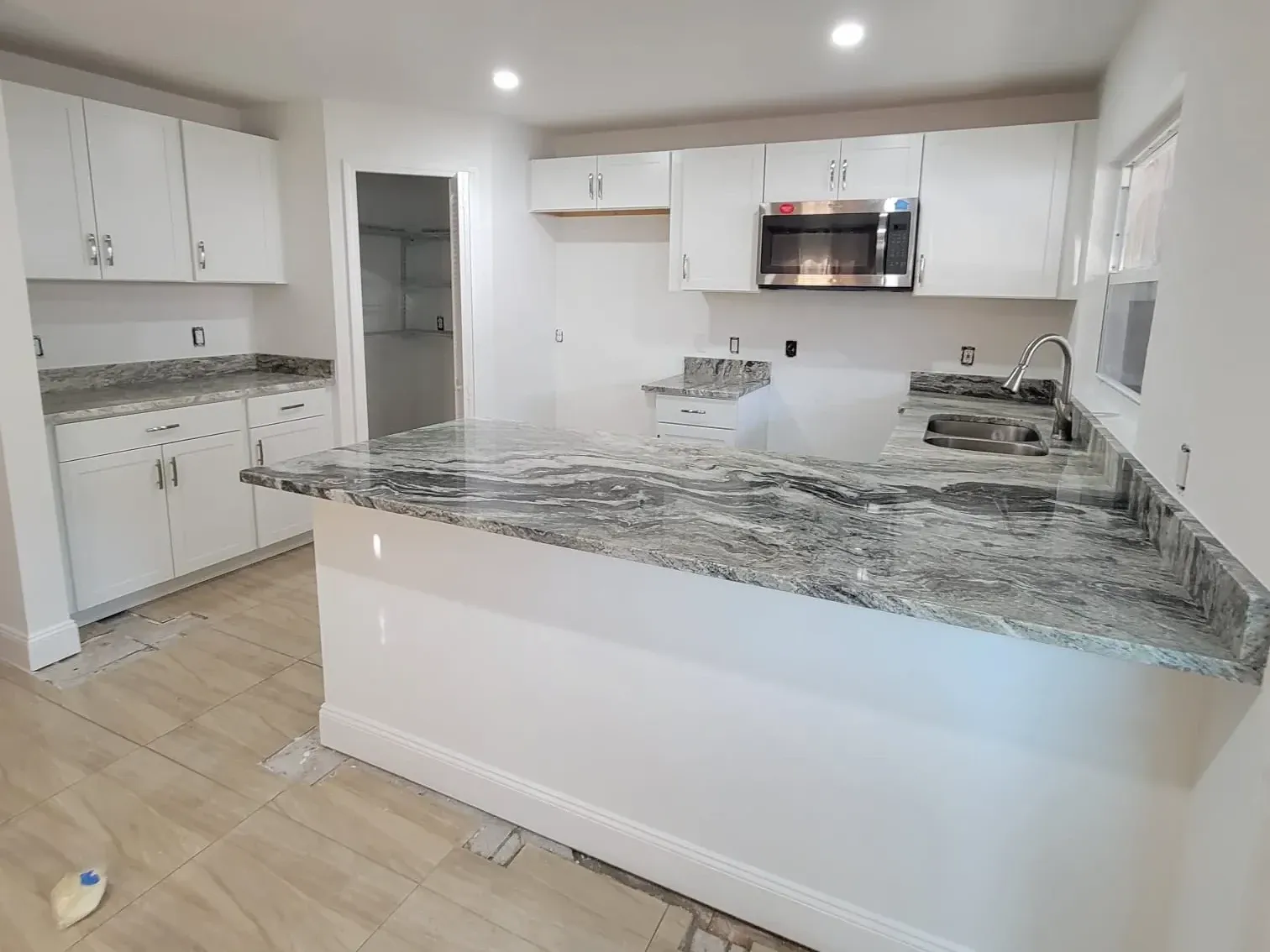 Open kitchen with white shaker cabinets, a gray-veined granite peninsula, stainless microwave and wood-look plank tile floor